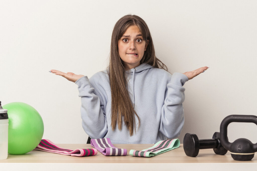 A young woman with long brown hair, wearing a light gray hoodie, sits at a table with various fitness equipment, including resistance bands, a green exercise ball, a water bottle, dumbbells, and a kettlebell. She raises her hands in a confused or uncertain gesture, biting her lip, as if unsure about which equipment to use. The background is a plain white wall.