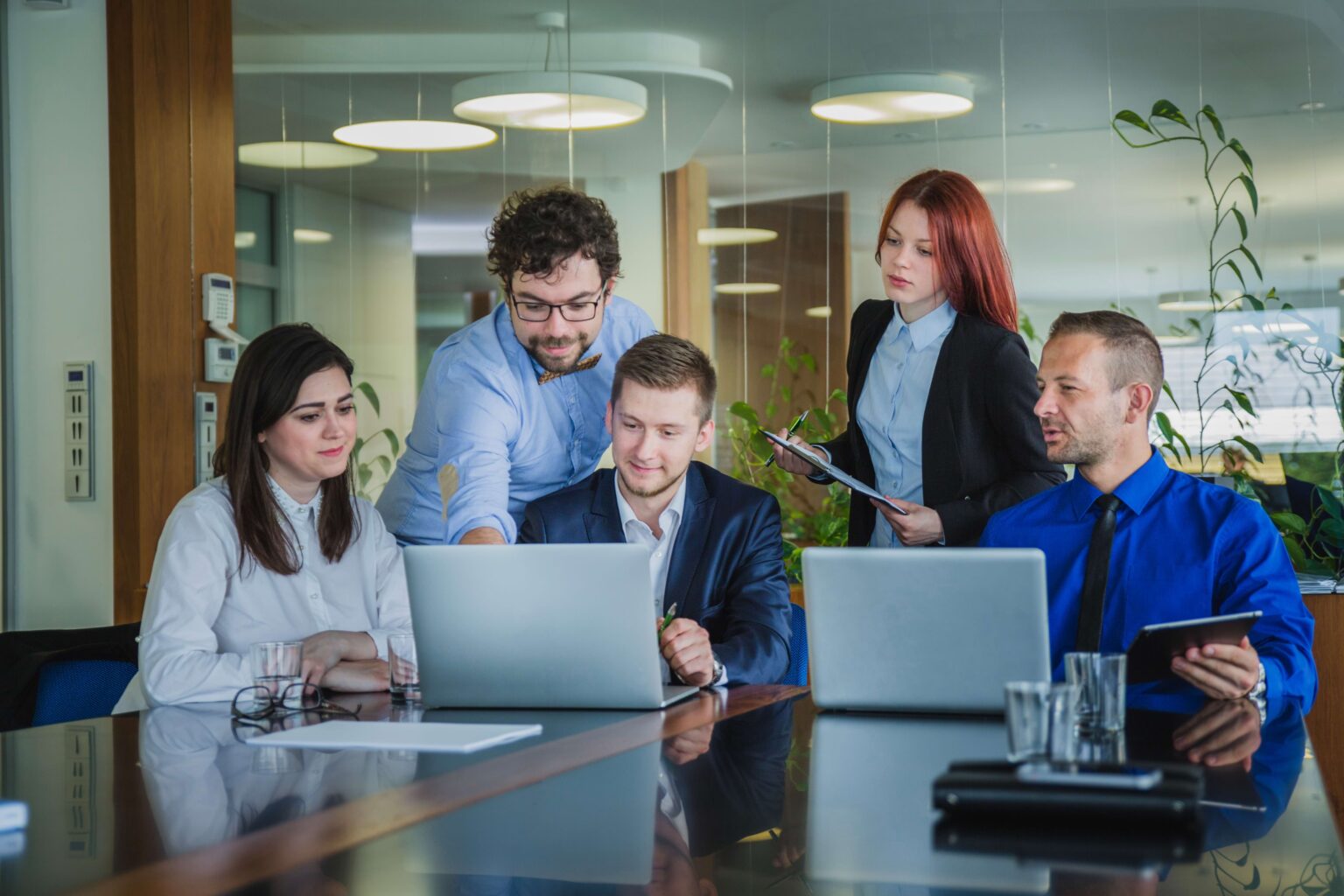 A group of five professionals in business attire collaborating in a modern office. Two men are using laptops, while a colleague in a blue shirt is explaining something. A woman with red hair is taking notes on a clipboard. The office features glass walls, greenery, and contemporary lighting.