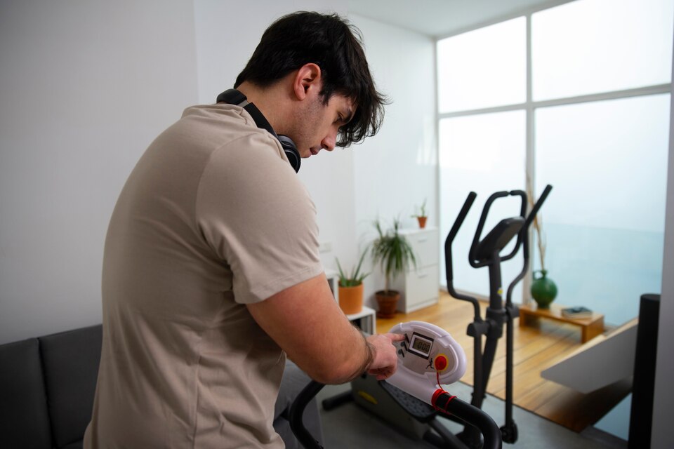 A young man with dark hair, wearing a beige t-shirt and black headphones around his neck, adjusts the settings on a stationary exercise bike in a modern, well-lit home. The room has large windows, potted plants, and minimalistic decor.