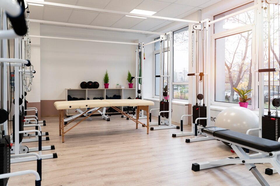 A well-lit physical rehabilitation therapy room equipped with exercise machines, resistance bands, and a massage table at the center. The space features wooden flooring, large windows, and organized shelves with fitness accessories and potted plants for a calming atmosphere.