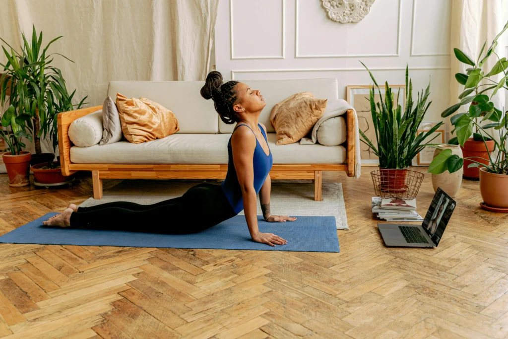 A young woman with braided hair practices yoga in a cozy living room, performing an upward-facing dog pose on a blue mat. She wears a blue top and black leggings, following an online session on a laptop, surrounded by plants and modern decor.